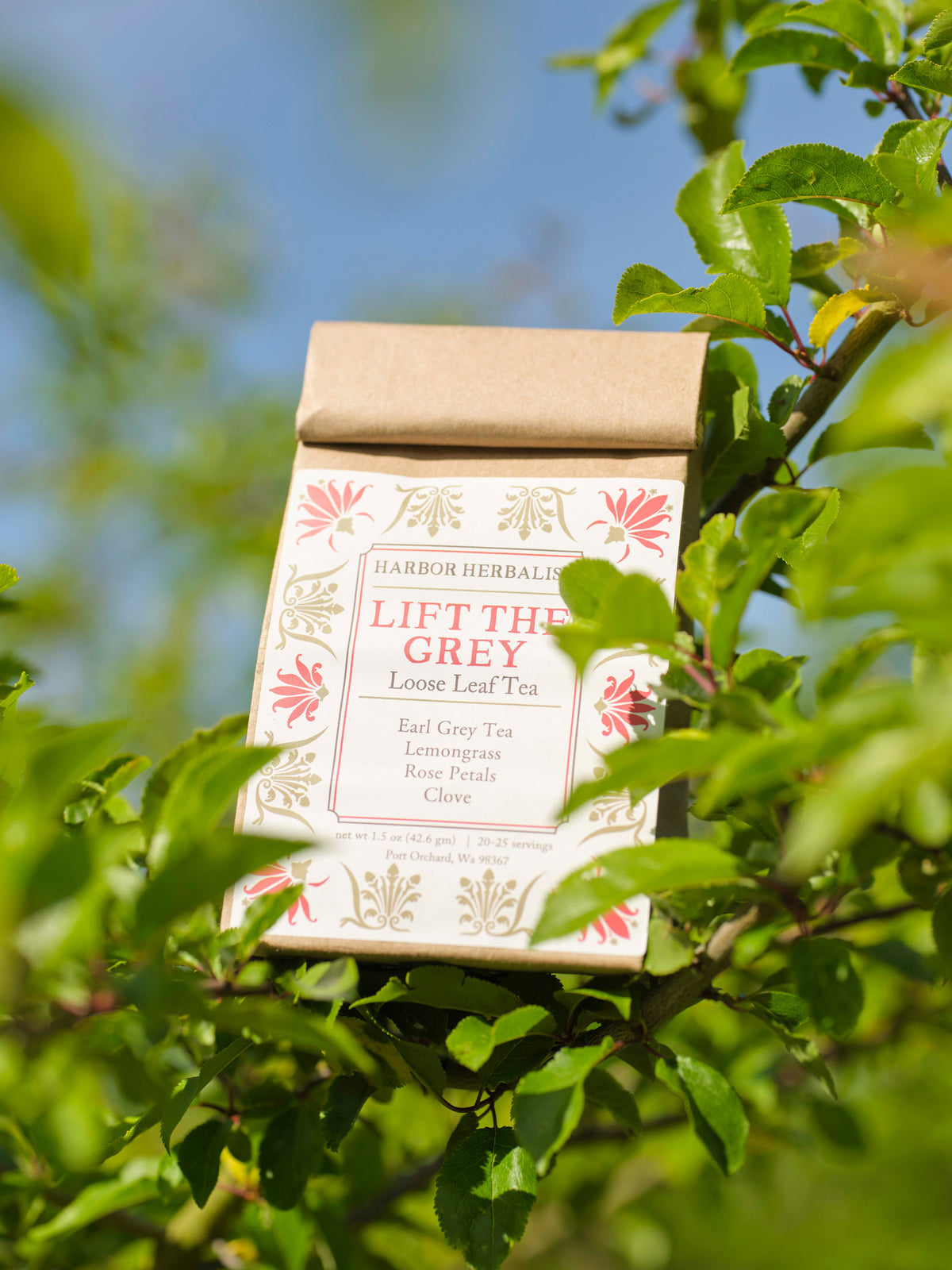 A kraft paper bag of Harbor Herbal 'Lift the Grey' loose leaf tea, featuring a white label with red and gold botanical illustrations, is shown hanging from a green leafy branch outdoors against a blue sky background.
