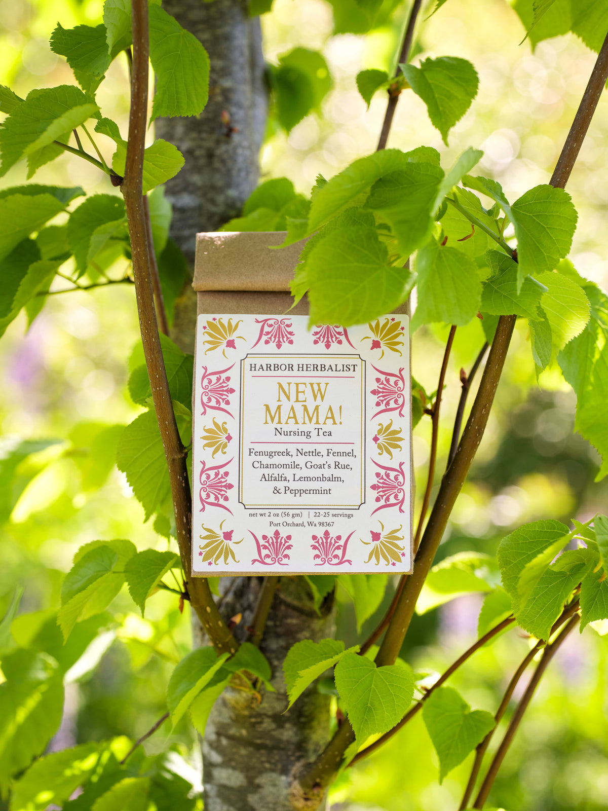 A bag of Harbor Herbalist 'New Mama' nursing tea hangs on a tree trunk surrounded by green leaves and sunlight. The front label features a white background with pink and yellow botanical illustrations and black text.