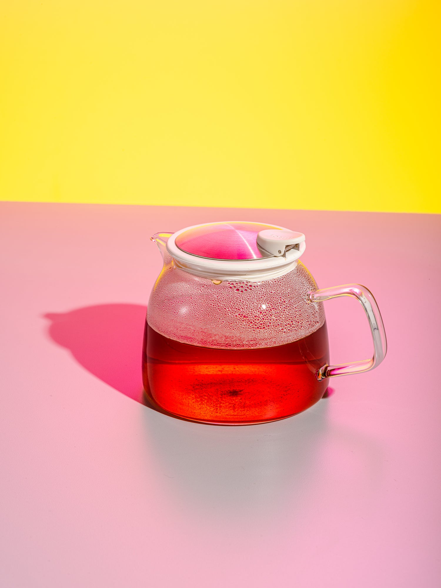 A clear, translucent teapot with brewed black tea sitting on a pink table with a yellow studio background.