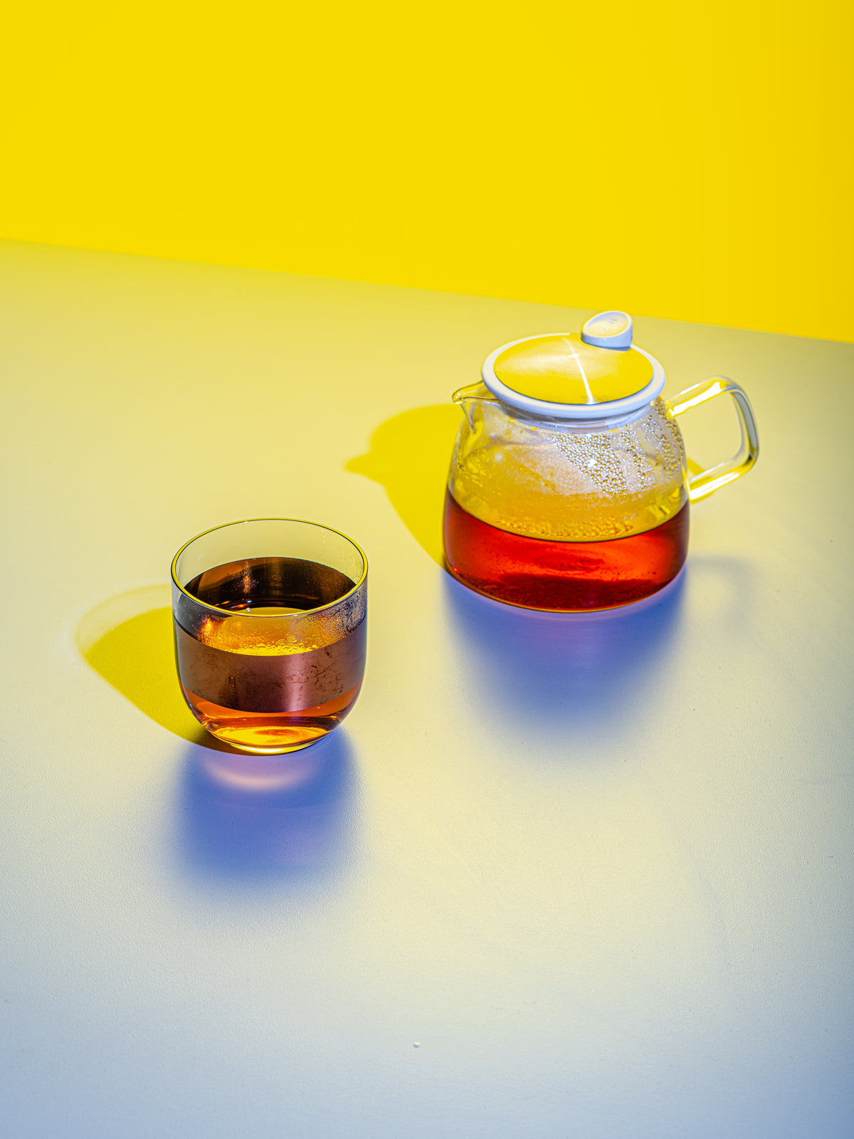 A cup of brewed black tea in a clear, translucent mug next to a clear, translucent teapot with more brewed tea inside.