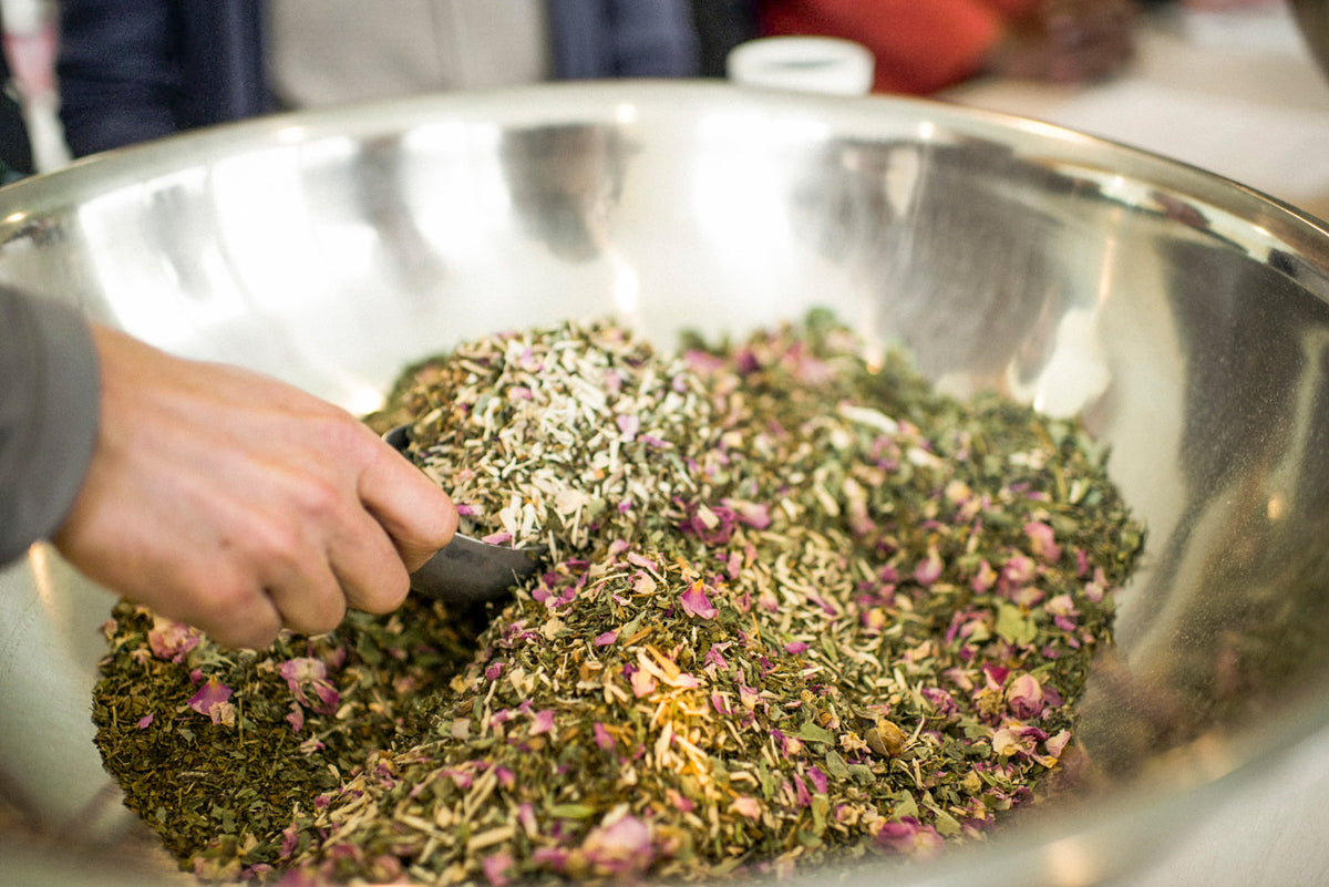 A close-up of a hand mixing a blend of loose-leaf tea, herbs, and pink flower petals inside a large stainless steel bowl.