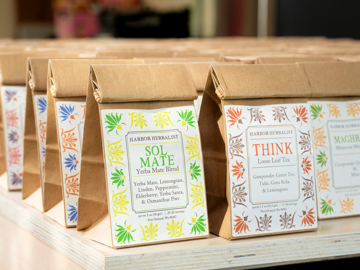 A row of kraft paper tea bags on a wooden shelf, each with a decorative label. The front bag is labeled 'SOL MATE Yerba Mate Blend' and the next visible bag is labeled 'THINK Loose Leaf Tea'.