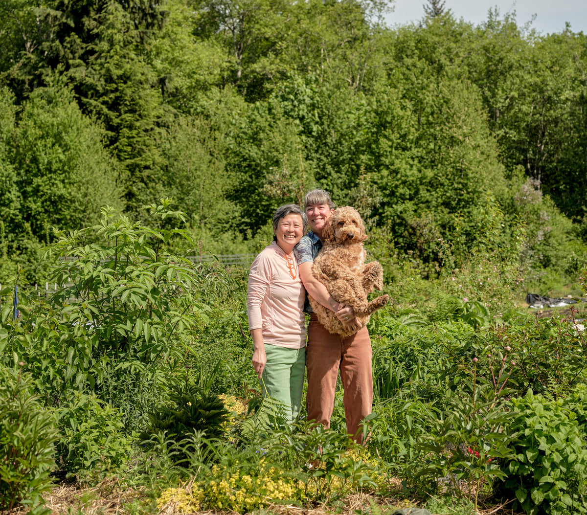 Two people standing in a lush green garden, with one holding a large brown dog. The person on the left wears a light pink top and green pants, while the person on the right wears a blue shirt and brown pants.