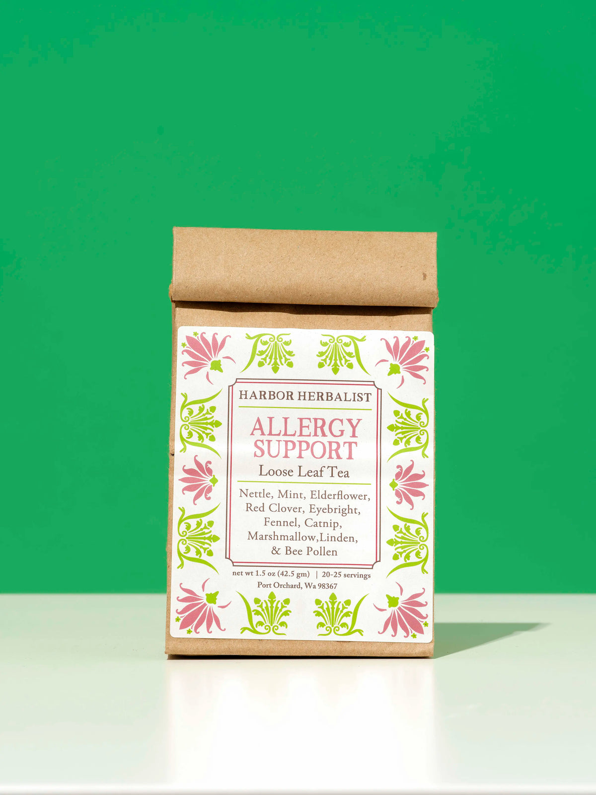 A kraft paper bag of Harbor Herbalist Allergy Support Loose Leaf Tea, featuring a white label with pink and green botanical illustrations, set against a solid green background.