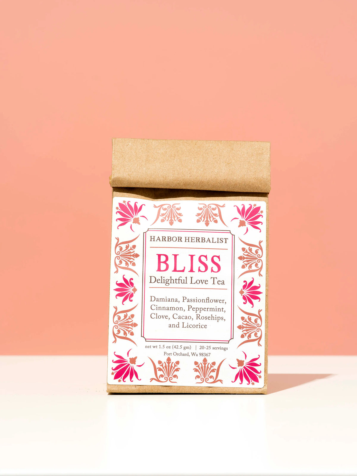 A kraft paper bag of Harbor Herbalist Bliss Delightful Love Tea, standing upright on a white surface against a soft peach-pink background. The packaging features a white front with red and pink botanical illustrations.