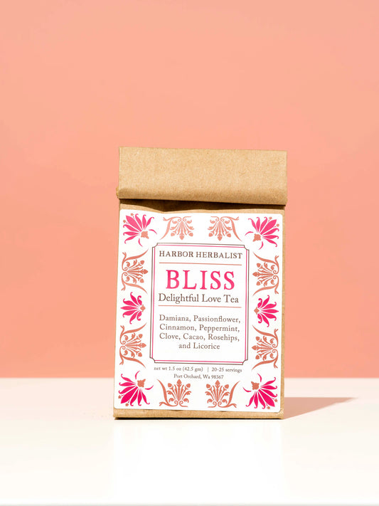 A kraft paper bag of Harbor Herbalist Bliss Delightful Love Tea, standing upright on a white surface against a soft peach-pink background. The packaging features a white front with red and pink botanical illustrations.