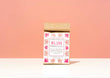 A kraft paper bag of Harbor Herbalist Bliss Delightful Love Tea, standing upright on a white surface against a soft peach-pink background. The packaging features a white front with red and pink botanical illustrations.