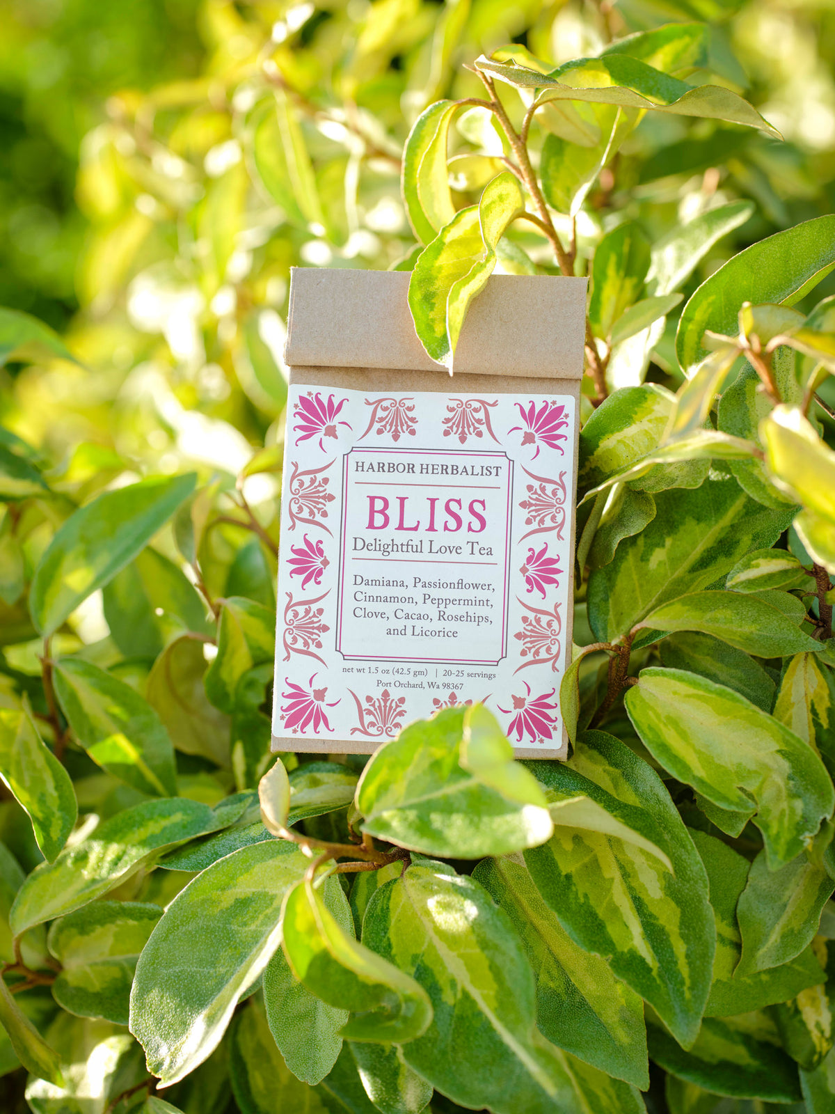 A kraft paper bag of Harbor Herbalist Bliss Delightful Love Tea stands upright among green variegated leaves outdoors in bright natural light. The packaging features a white front with red and pink botanical illustrations.