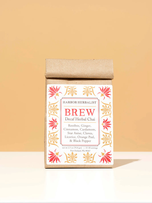 A kraft paper bag of Harbor Herbalist Brew Decaf Herbal Chai tea stands centered on a white surface against a warm beige background. The packaging features red and gold botanical illustrations.