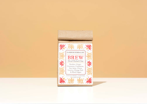 A kraft paper bag of Harbor Herbalist Brew Decaf Herbal Chai tea stands centered on a white surface against a warm beige background. The packaging features red and gold botanical illustrations.