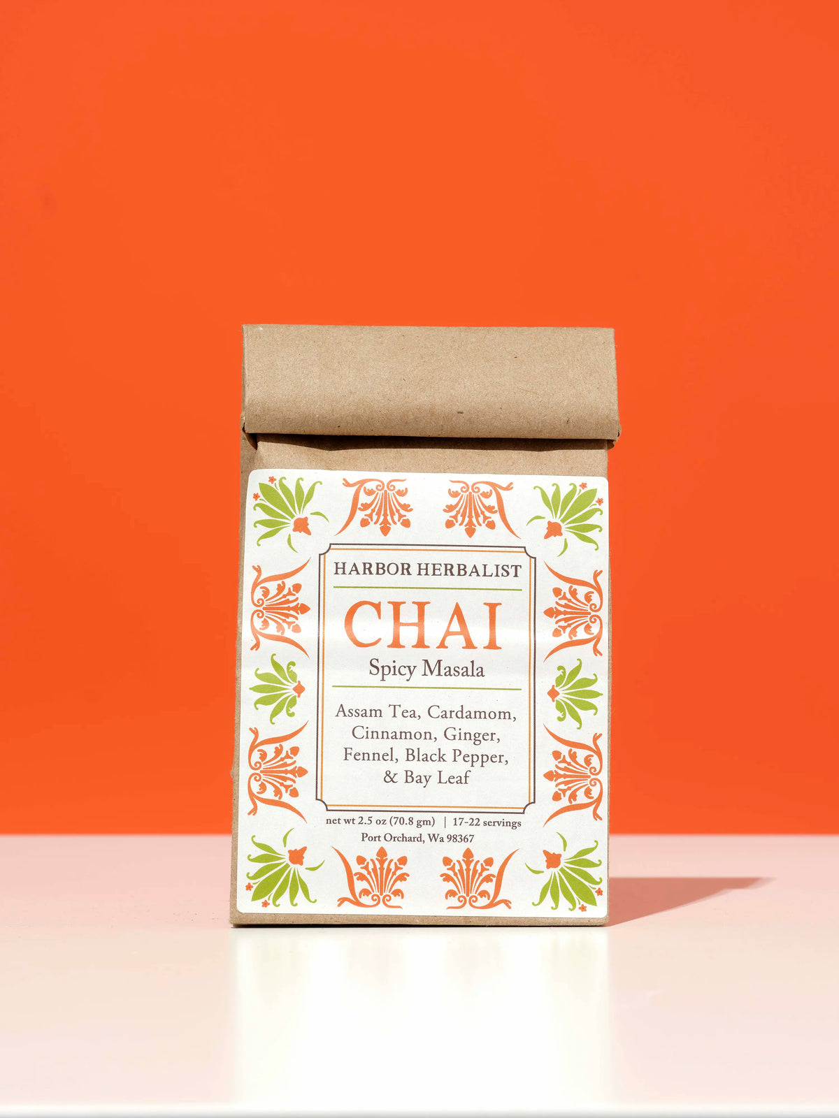 A kraft paper bag of Harbor Herbalist Chai Spicy Masala tea stands upright on a white surface against a solid orange background. The packaging features a white front label with orange and green botanical illustrations.