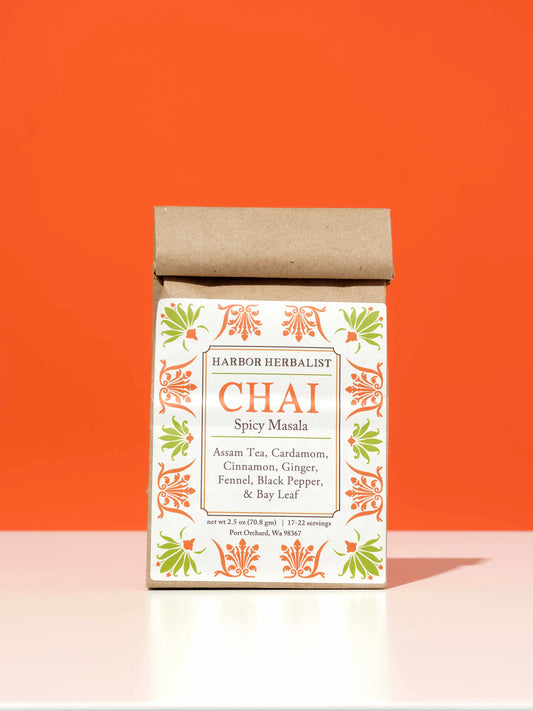 A kraft paper bag of Harbor Herbalist Chai Spicy Masala tea stands upright on a white surface against a solid orange background. The packaging features a white front label with orange and green botanical illustrations.