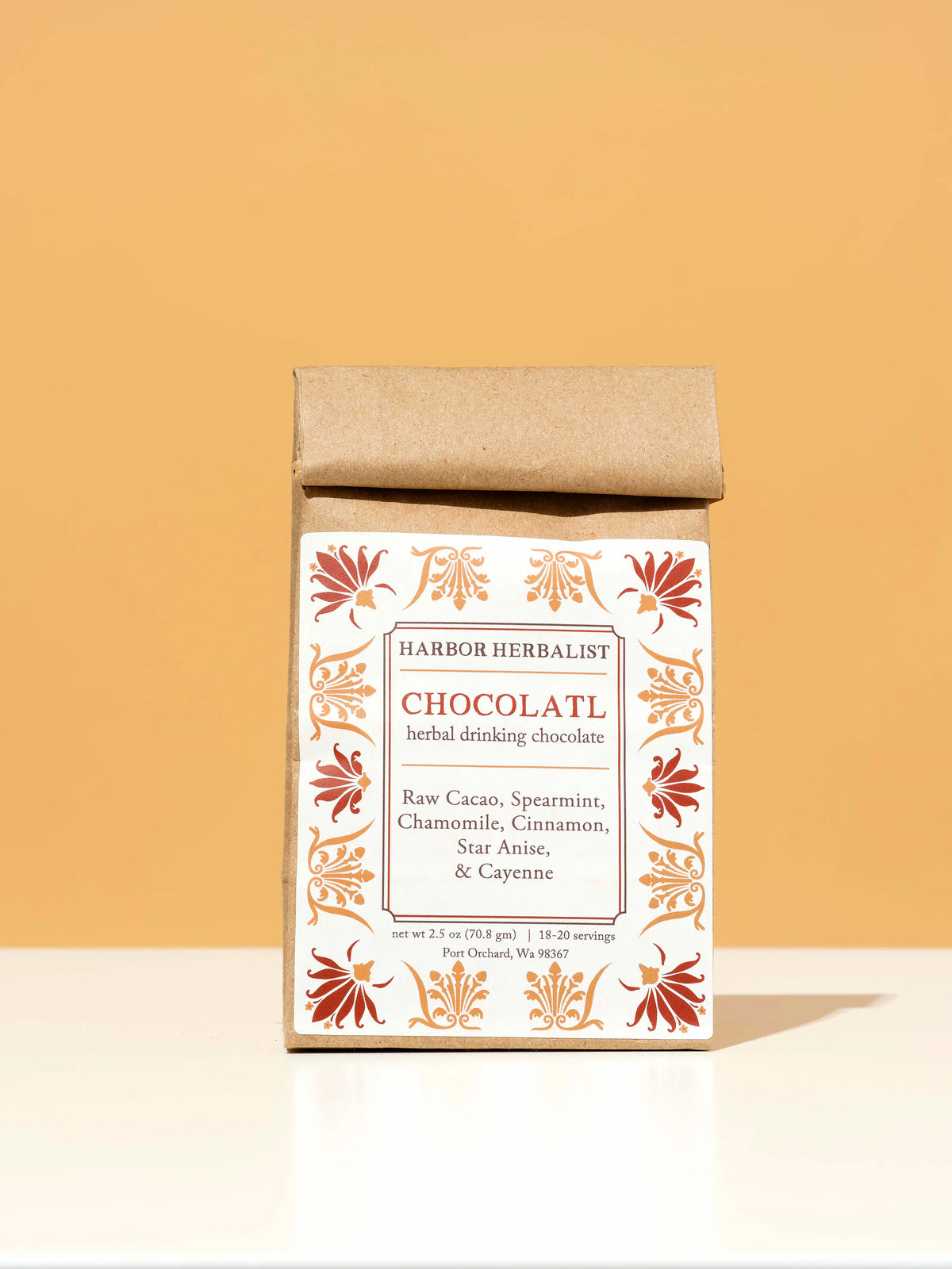 A kraft paper bag for Harbor Herbalist Chocolat, featuring a white label decorated with red and orange botanical illustrations, standing upright on a white surface against a warm beige background.
