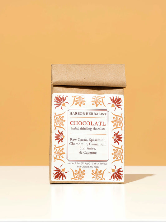 A kraft paper bag for Harbor Herbalist Chocolat, featuring a white label decorated with red and orange botanical illustrations, standing upright on a white surface against a warm beige background.