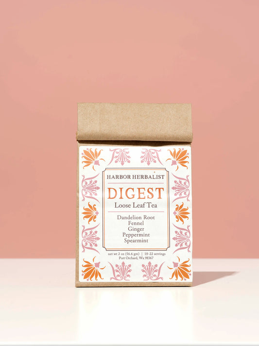 A kraft paper bag of Harbor Herbalist Digest Loose Leaf Tea stands upright on a white surface against a solid pink background. The packaging features a white label decorated with orange and pink botanical illustrations.