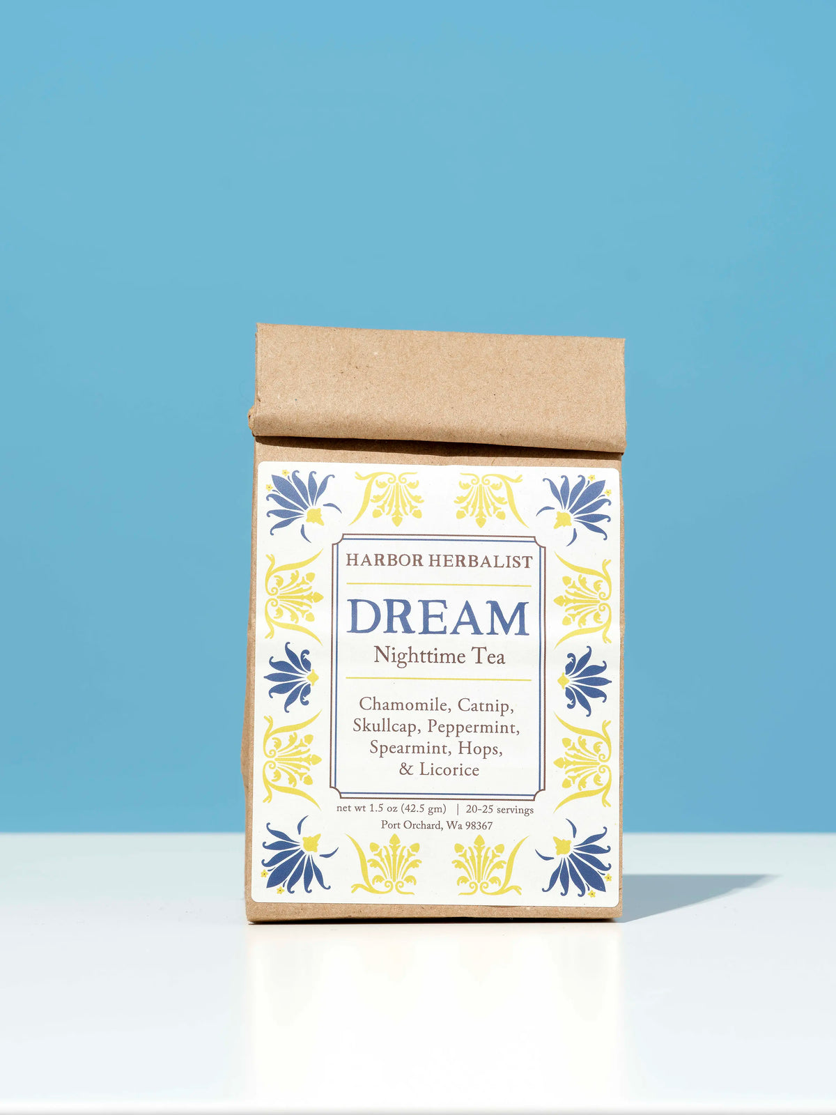A kraft paper bag of Harbor Herbalist Dream Nighttime Tea stands upright on a white surface against a solid blue background. The packaging features a white front label with blue and yellow botanical illustrations.