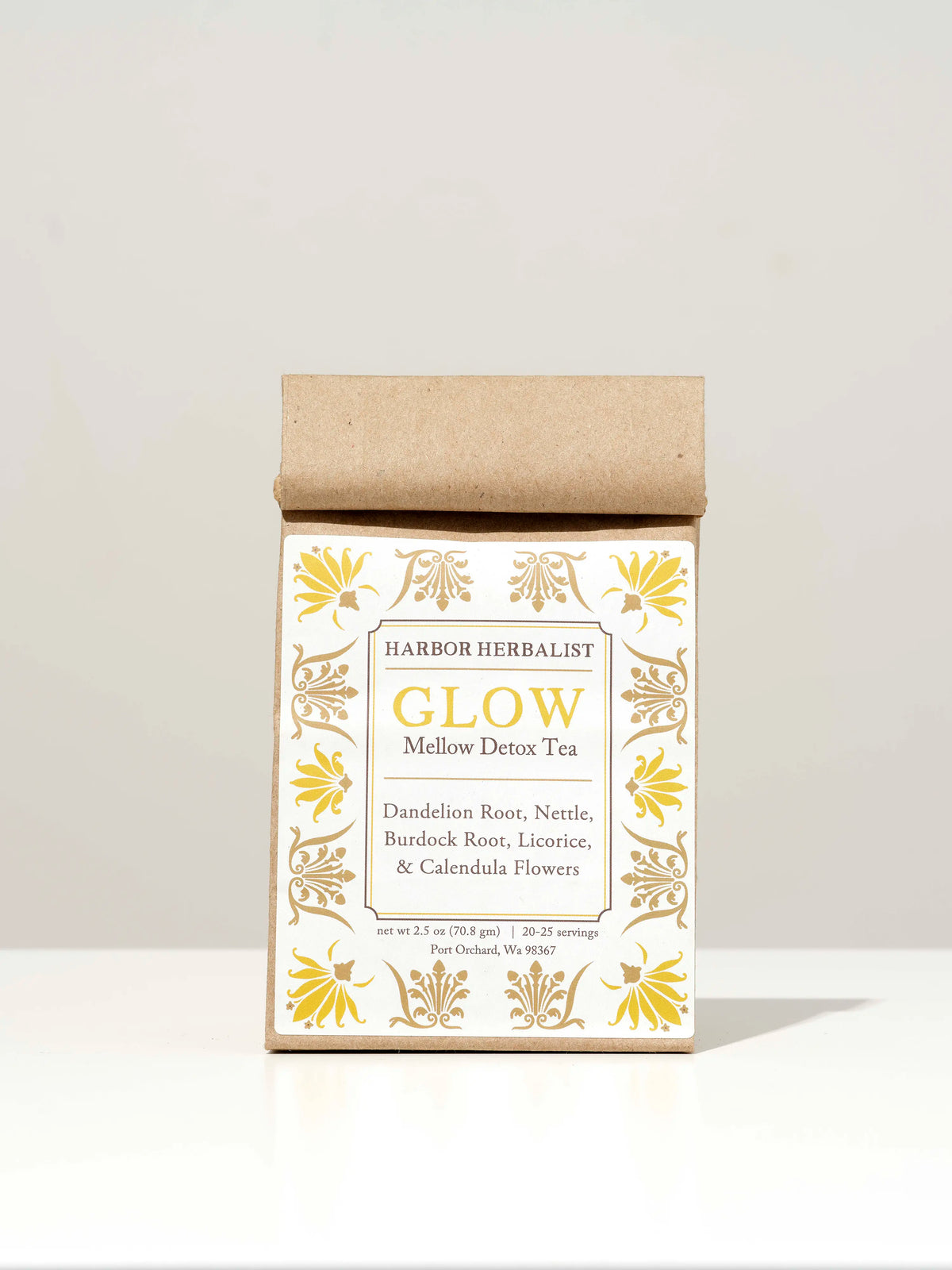 A kraft paper bag of Harbor Herbalist Glow Loose Leaf Tea, featuring a white label with yellow and tan botanical illustrations, set against a solid grey background.