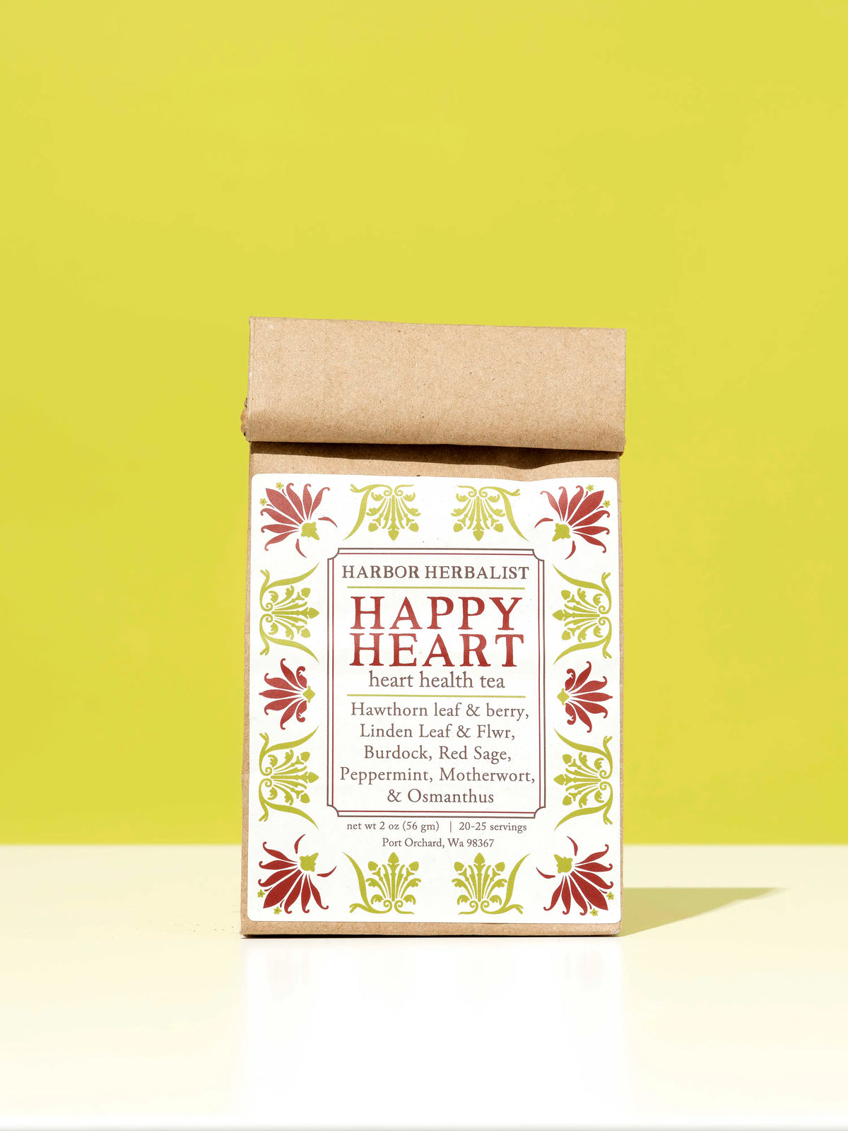A kraft paper bag of Harbor Herbalist Happy Heart heart health tea stands upright on a white surface against a bright yellow-green background. The packaging features a white front label with red and green botanical illustrations.