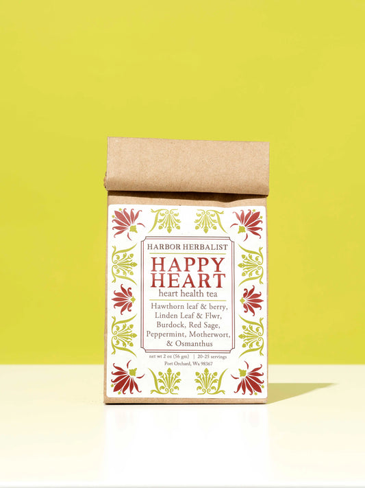 A kraft paper bag of Harbor Herbalist Happy Heart heart health tea stands upright on a white surface against a bright yellow-green background. The packaging features a white front label with red and green botanical illustrations.
