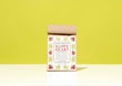 A kraft paper bag of Harbor Herbalist Happy Heart heart health tea stands upright on a white surface against a bright yellow-green background. The packaging features a white front label with red and green botanical illustrations.
