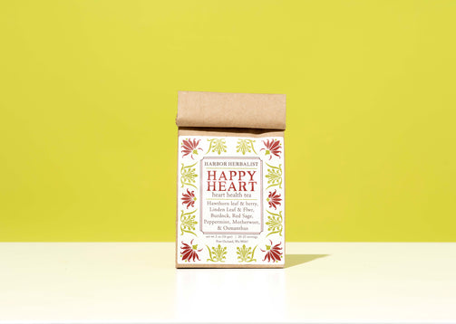 A kraft paper bag of Harbor Herbalist Happy Heart heart health tea stands upright on a white surface against a bright yellow-green background. The packaging features a white front label with red and green botanical illustrations.