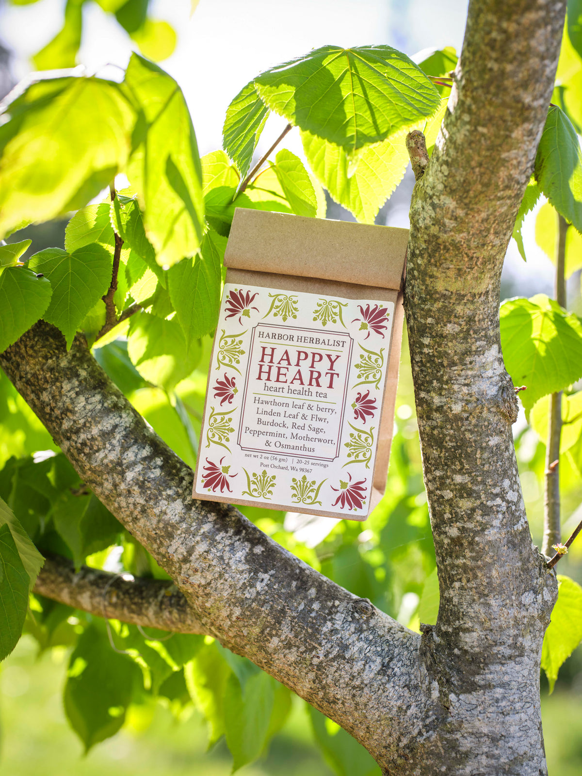 A kraft paper bag of Happy Heart heart health tea hangs from a tree branch outdoors, surrounded by green leaves and natural sunlight. The packaging features a white front label with red and green botanical illustrations.