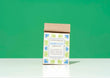 A kraft paper bag of Harbor Herbalist Hercules Athlete's Tea stands upright on a white surface against a solid green background. The packaging features a white front label decorated with blue and green botanical illustrations.