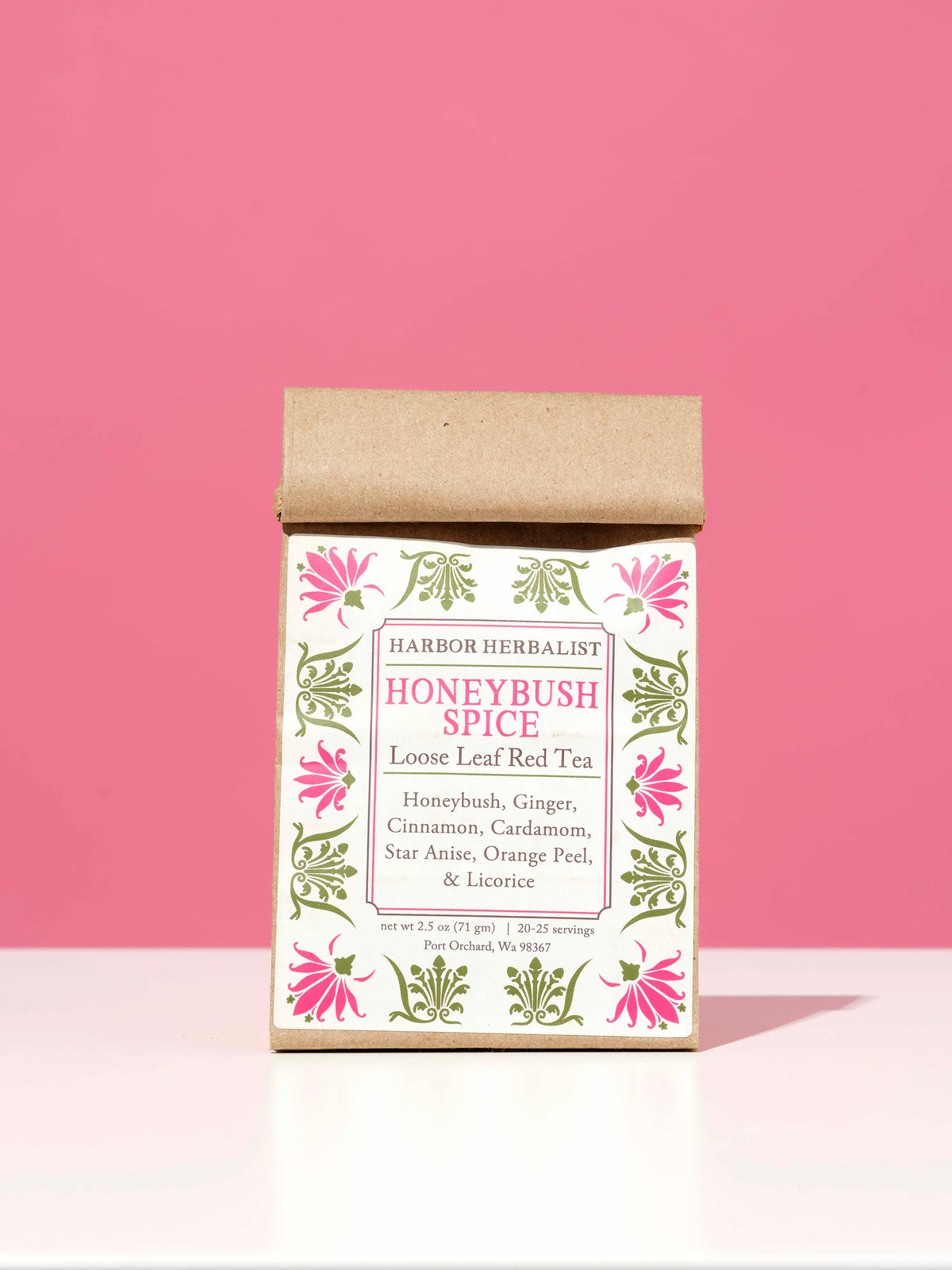 A kraft paper bag of Harbor Herbalist Honeybush Spice loose leaf tea stands centered on a white surface against a solid pink background. The bag features a white label decorated with pink and green botanical illustrations.