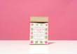 A kraft paper bag of Harbor Herbalist Honeybush Spice loose leaf tea stands centered on a white surface against a solid pink background. The bag features a white label decorated with pink and green botanical illustrations.