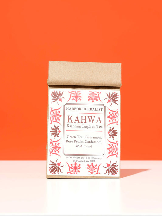 A kraft paper bag of Harbor Herbalist Kahwa Kashmiri Inspired Tea stands upright on a white surface against a solid orange background. The bag features a white front label with red botanical illustrations and black text.
