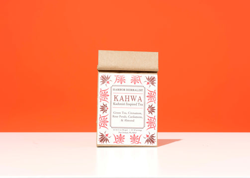 A kraft paper bag of Harbor Herbalist Kahwa Kashmiri Inspired Tea stands upright on a white surface against a solid orange background. The bag features a white front label with red botanical illustrations and black text.