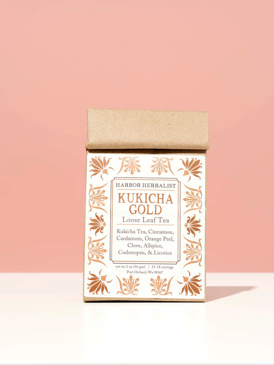 A kraft paper bag of Harbor Herbalist Kukicha Gold Loose Leaf Tea, featuring a white label and copper-gold botanical illustrations, set against a solid pink background on a white surface.
