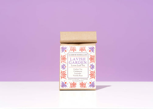 A bag of Harbor Herbalist Lavish Garden loose leaf herbal tea, featuring a white label decorated with red and purple botanical illustrations, set against a solid lavender background on a white surface.