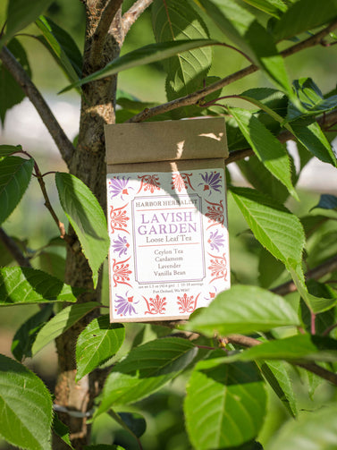 A bag of Harbor Herbalist Lavish Garden loose leaf herbal tea, hanging from a tree with leaves, featuring a white label decorated with red and purple botanical illustrations.