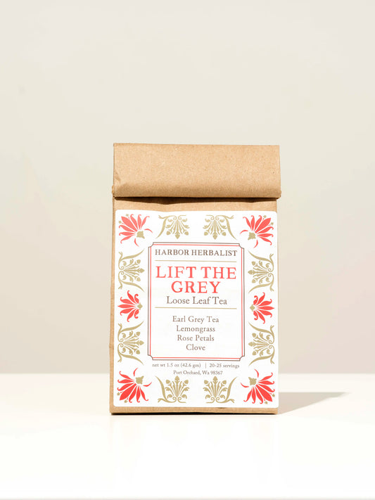 A kraft paper bag of Harbor Herbalist Lift The Grey Loose Leaf Tea, featuring a white label with red and gold botanical illustrations, set against a plain off-white background.
