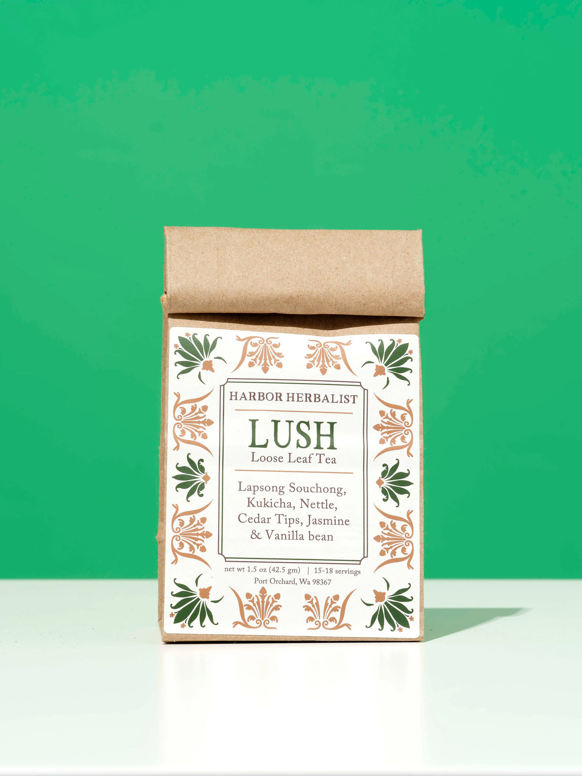 A bag of Harbor Herbalist Lush loose leaf tea, featuring a white label decorated with green and gold botanical illustrations, stands upright on a white surface against a bright green background.