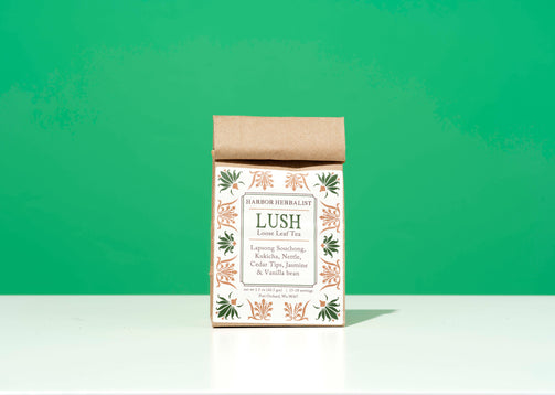 A bag of Harbor Herbalist Lush loose leaf tea, featuring a white label decorated with green and gold botanical illustrations, stands upright on a white surface against a bright green background.