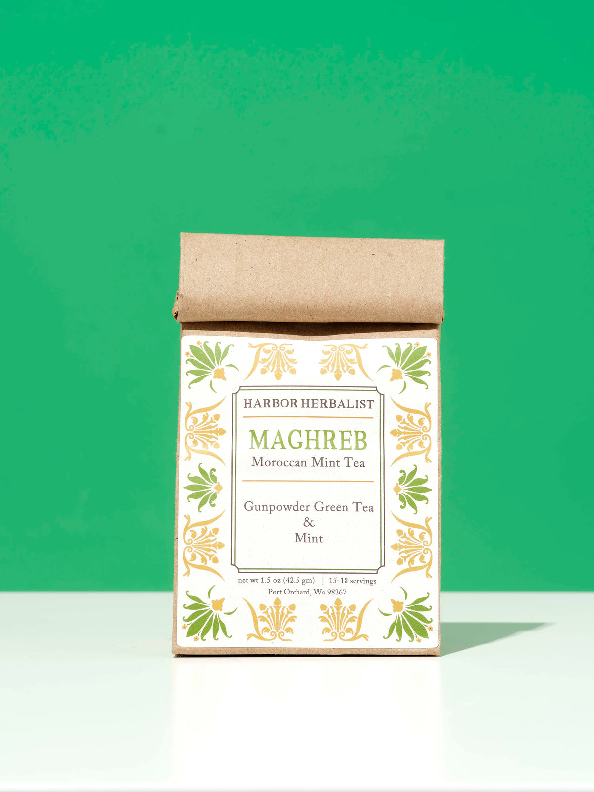 A bag of Harbor Herbalist Maghreb Moroccan Mint loose leaf tea, featuring a white label decorated with green and yellow botanical illustrations, stands on a white surface against a vibrant green background.
