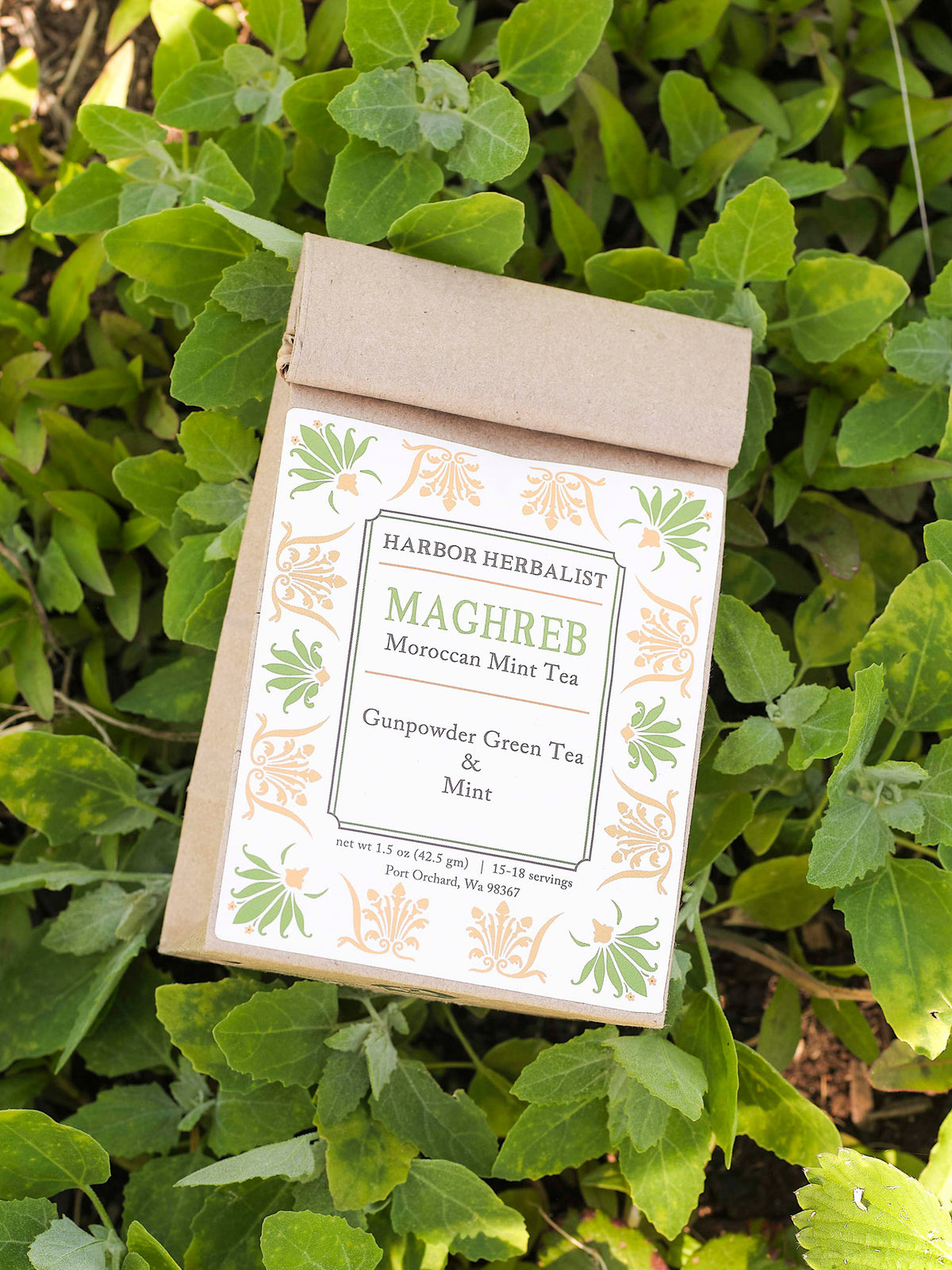 A paper bag of Harbor Herbalist Maghreb Moroccan Mint Tea lies on a bed of green leaves. The front of the bag features a white label with green and gold botanical illustrations and text identifying the product as Gunpowder Green Tea & Mint.