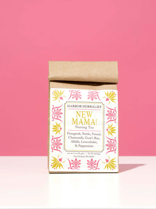 A small kraft paper bag of Harbor Herbalist 'New Mama! Nursing Tea' stands upright on a white surface against a solid pink background. The front label features a white background with pink and yellow botanical illustrations and black text.