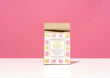 A small kraft paper bag of Harbor Herbalist 'New Mama! Nursing Tea' stands upright on a white surface against a solid pink background. The front label features a white background with pink and yellow botanical illustrations and black text.
