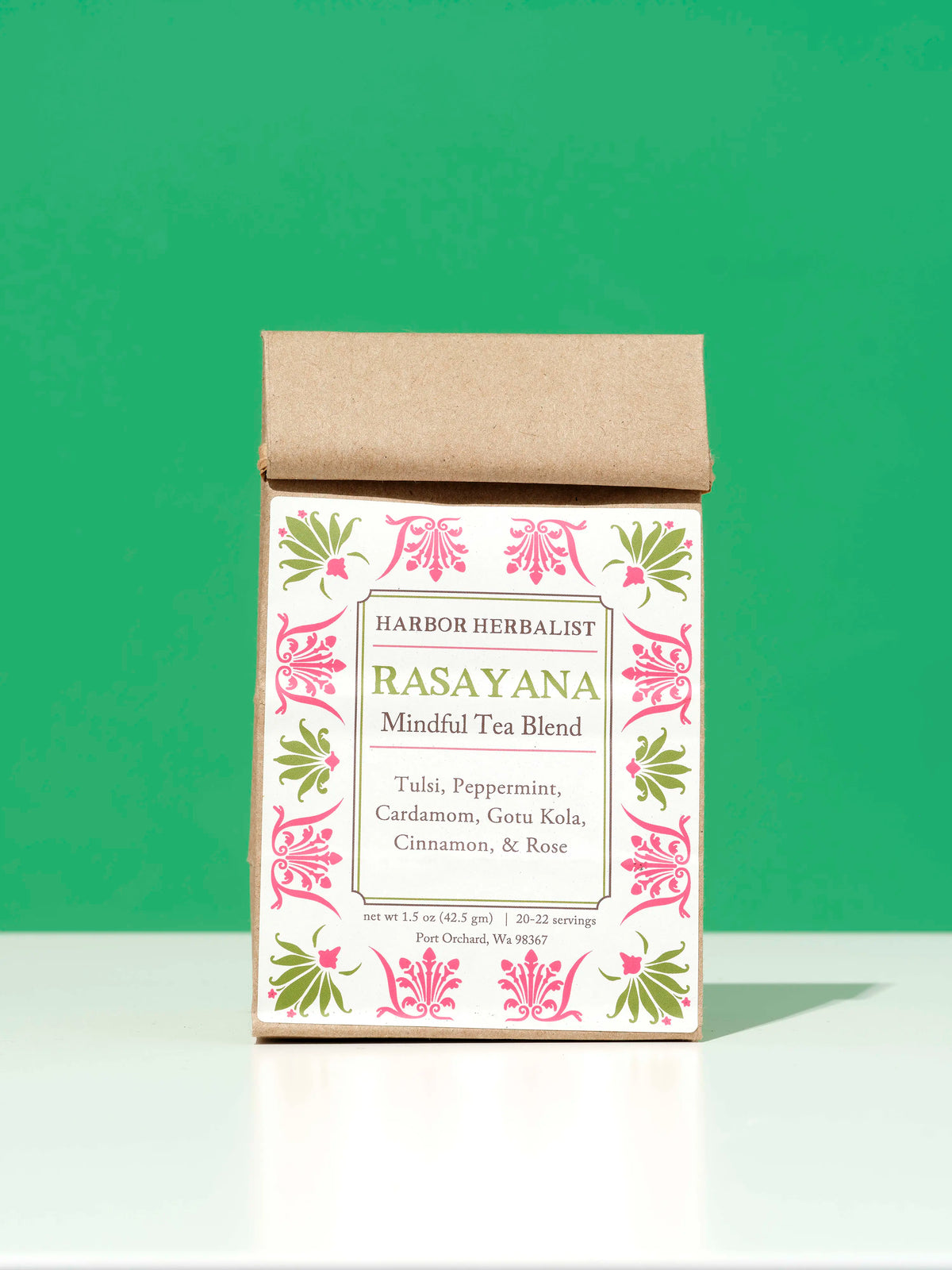A bag of Harbor Herbalist Rasayana Mindful Tea Blend, featuring a kraft paper top and a white front label with pink and green botanical illustrations, set against a solid green background.