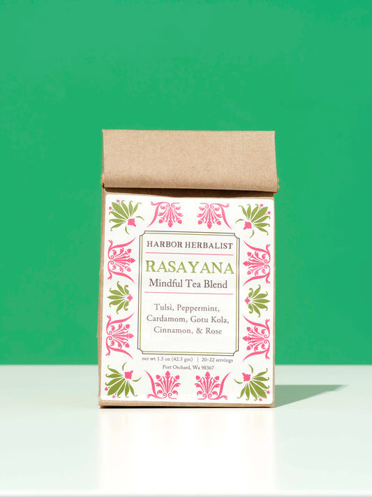 A bag of Harbor Herbalist Rasayana Mindful Tea Blend, featuring a kraft paper top and a white front label with pink and green botanical illustrations, set against a solid green background.
