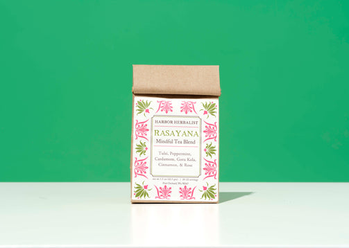 A bag of Harbor Herbalist Rasayana Mindful Tea Blend, featuring a kraft paper top and a white front label with pink and green botanical illustrations, set against a solid green background.