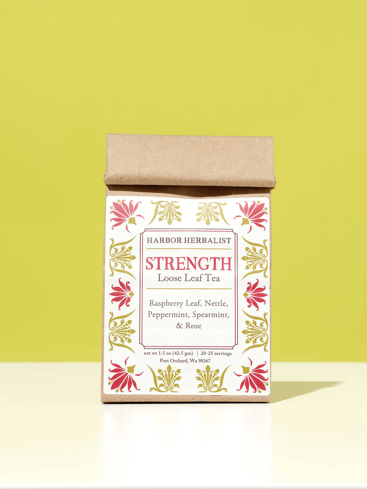 A kraft paper bag of Harbor Herbalist Strength Loose Leaf Tea stands upright on a white surface against a solid yellow-green background. The kraft paper box has a white label decorated with pink and gold botanical illustrations.