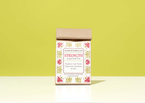 A kraft paper bag of Harbor Herbalist Strength Loose Leaf Tea stands upright on a white surface against a solid yellow-green background. The kraft paper box has a white label decorated with pink and gold botanical illustrations.