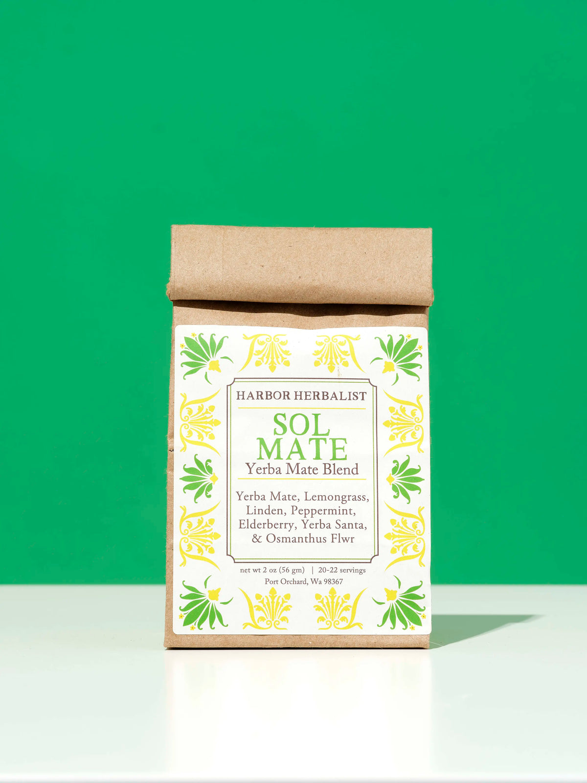 A kraft paper bag of Harbor Herbalist Sol Mate Yerba Mate blend tea with a white label with yellow and green botanical illustrations, set against a solid green background on a white surface.