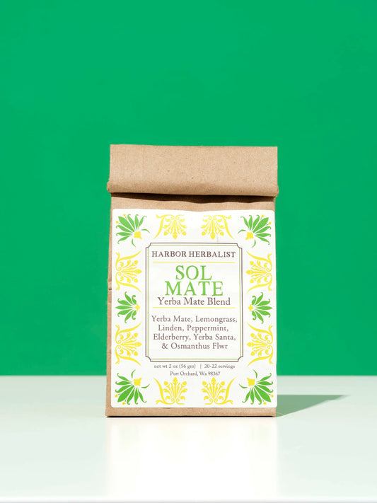 A kraft paper bag of Harbor Herbalist Sol Mate Yerba Mate blend tea with a white label with yellow and green botanical illustrations, set against a solid green background on a white surface.