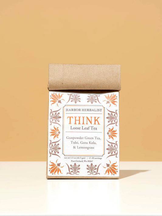 A kraft paper bag of Harbor Herbalist Think Loose Leaf Tea stands upright on a white surface against a warm beige background. The packaging features a white front label with orange and brown botanical illustrations.
