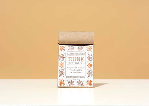 A kraft paper bag of Harbor Herbalist Think Loose Leaf Tea stands upright on a white surface against a warm beige background. The packaging features a white front label with orange and brown botanical illustrations.